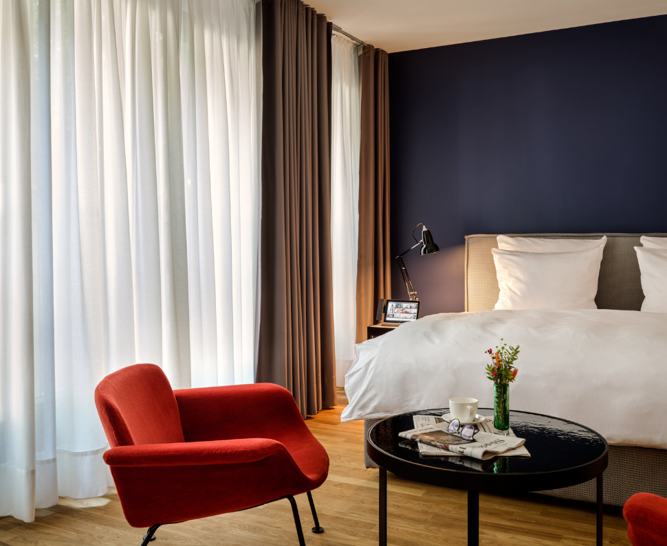 Stilvolles Schlafzimmer mit einem roten Stuhl, einer marineblauen Akzentwand und weißer Bettwäsche. Auf einem runden Tisch stehen eine Kaffeetasse und Blumen, während hohe Vorhänge das Licht sanft filtern.
