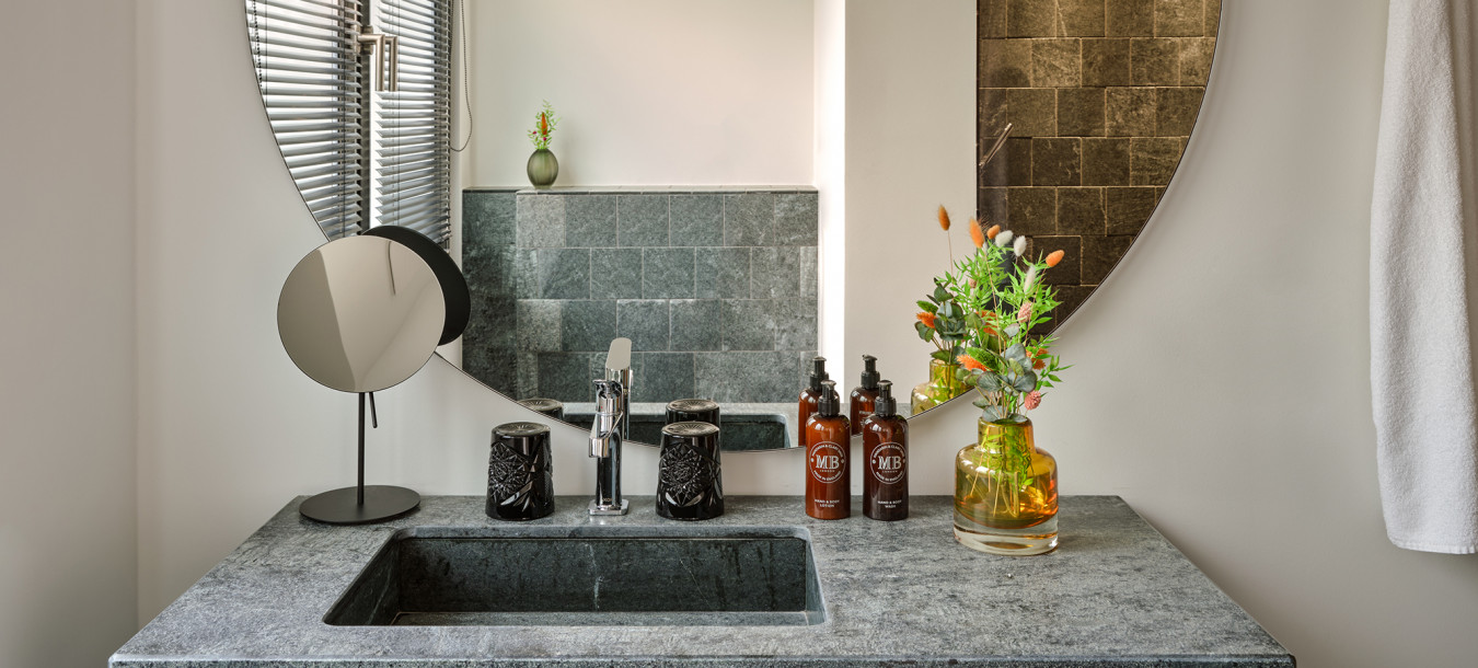 Modern bathroom vanity with a large round mirror, stone sink, toiletries, flowers in a vase, and a towel on the wall.