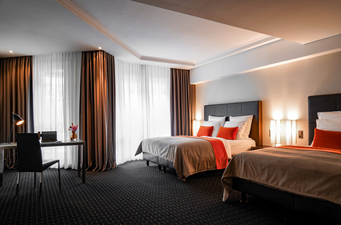 Modern hotel room with two double beds, a desk and chair, floor-to-ceiling windows, and warm lighting.