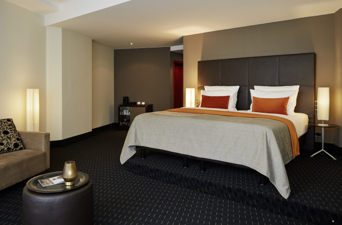 A modern hotel room with a large bed, orange and white pillows, soft lighting, and a sitting area with a small table.