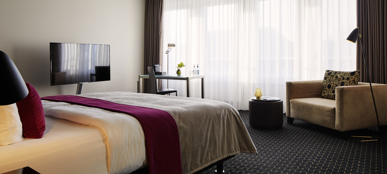 Modern hotel room with a large bed, TV, desk, armchair, floor lamp, and large windows covered with sheer curtains.