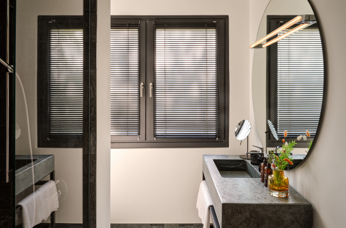 Modern bathroom with a round mirror, a gray countertop, black sink, towels, and window with closed blinds.