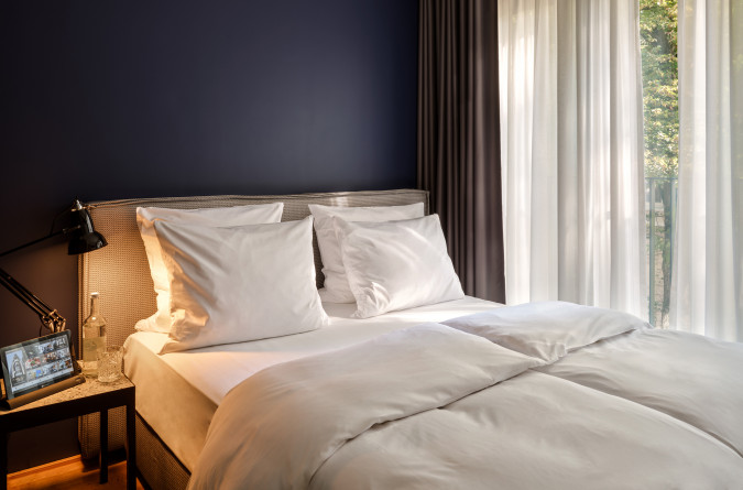 A neatly made bed with white linens near a window with sheer curtains, letting in natural light, in a modern bedroom.