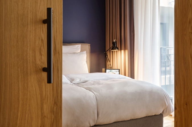 A cozy modern bedroom with a neatly made bed, wooden floor, dark blue accent wall, and soft natural light from a window.