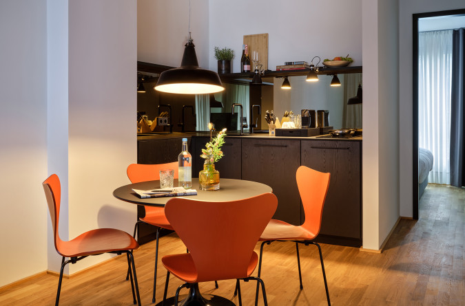 Modern kitchen and dining area with orange chairs, round table, hanging light, and an open view into a cozy bedroom.
