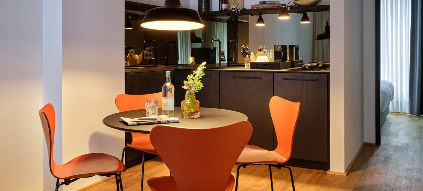 Modern dining area with round table, four orange chairs, and a kitchen in the background with dark cabinets and warm lighting.