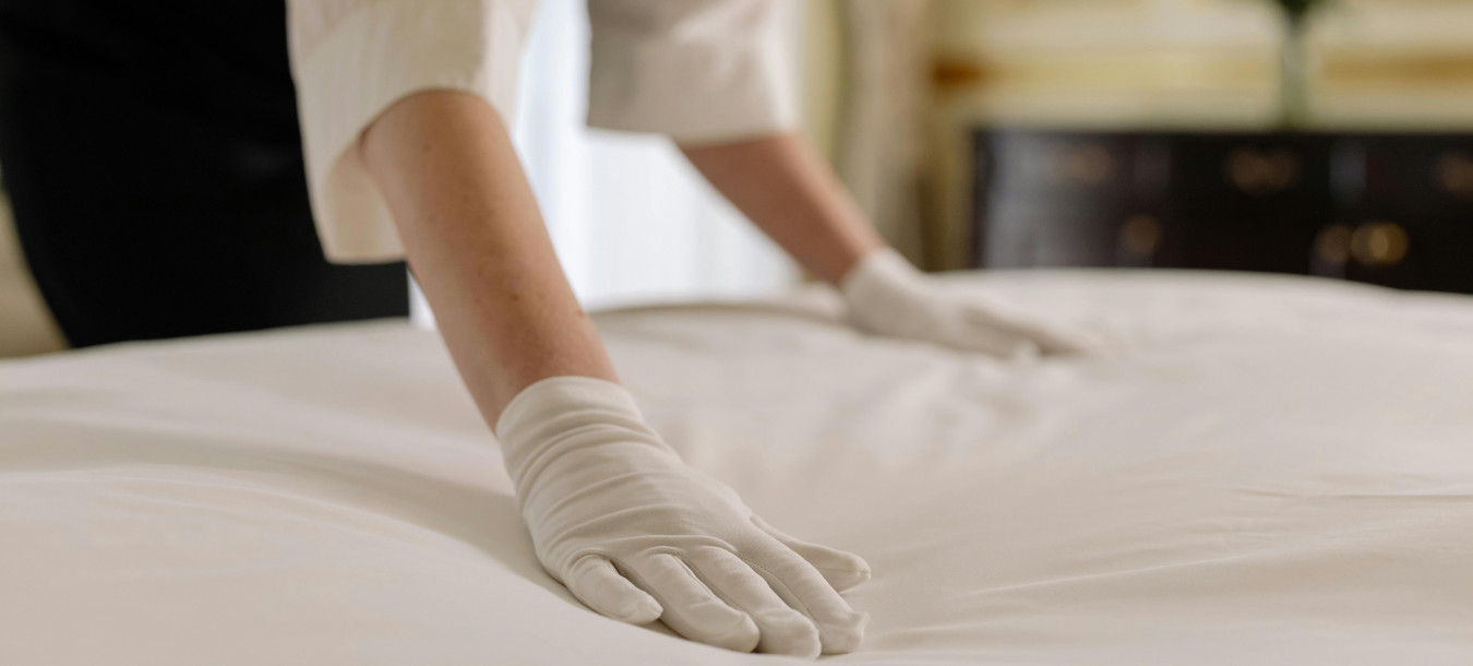 Eine Person in Uniform und weißen Handschuhen macht ein Bett und glättet die weißen Laken in einem gut beleuchteten Raum.